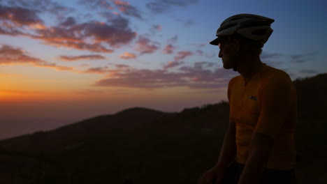 A-man-standing-on-a-bicycle-atop-the-mountain-gazes-at-the-sunset-while-the-camera-glides-on-a-Steadicam